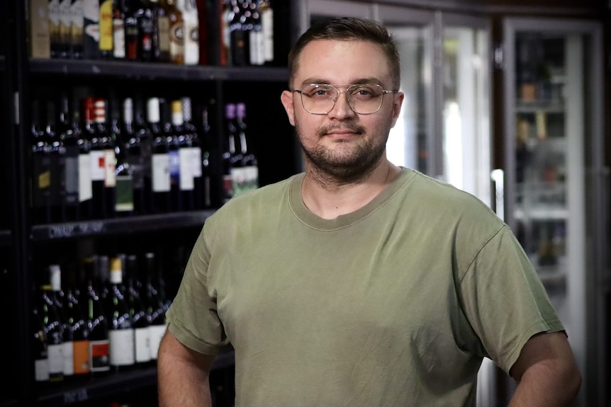 Man with glasses and short hair with t-shirt on smiles at camera in front of wine bottles