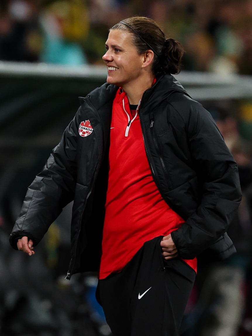 A woman in a red shirt and black jacket with brown hair smiles out of frame