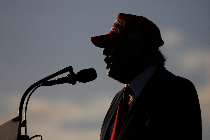 Donald Trump silloutted against a dark sky wearing a red cap and speaking into a microphone.
