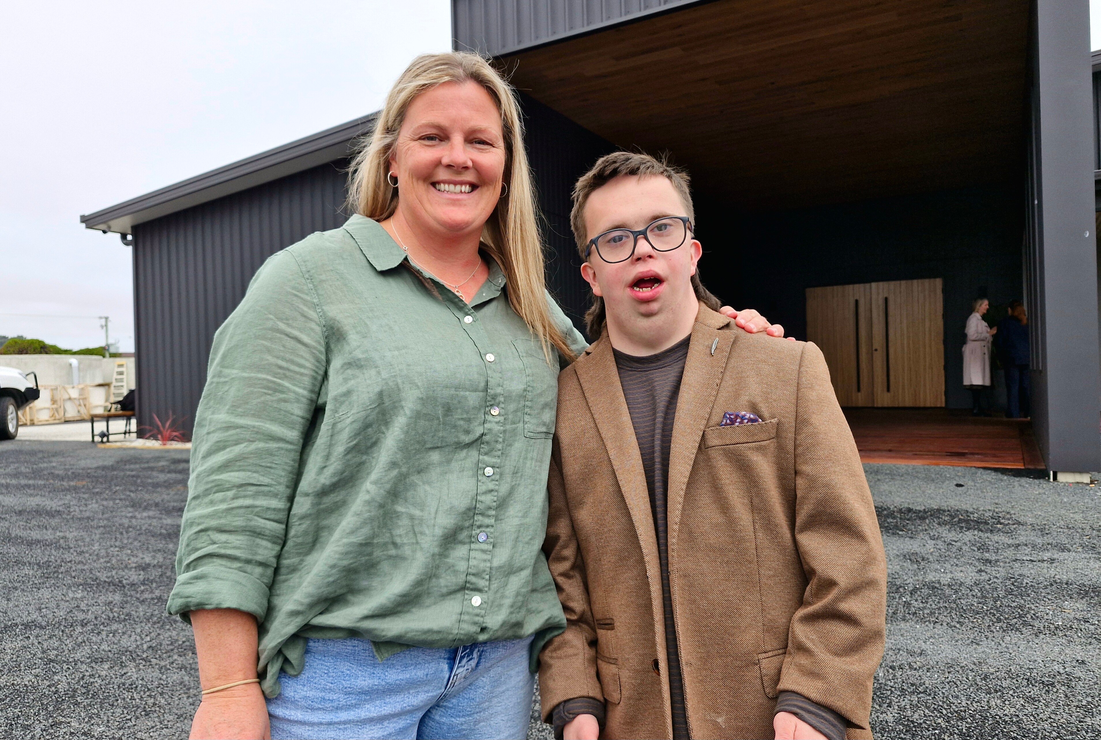 A blonde woman and a young man with down sydrome stand centre frame, smiling at the camera.