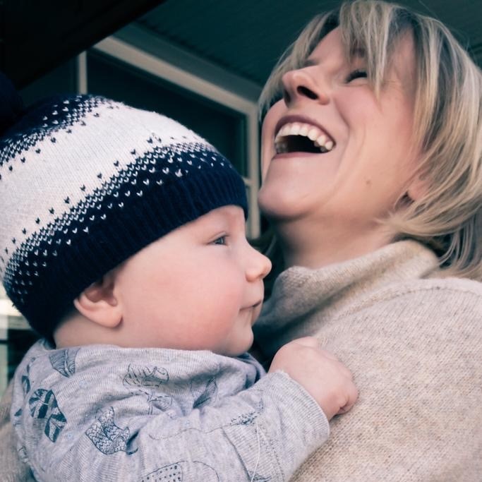 A mother throws her head back, laughing, while her baby smiles. He's wearing a beanie.