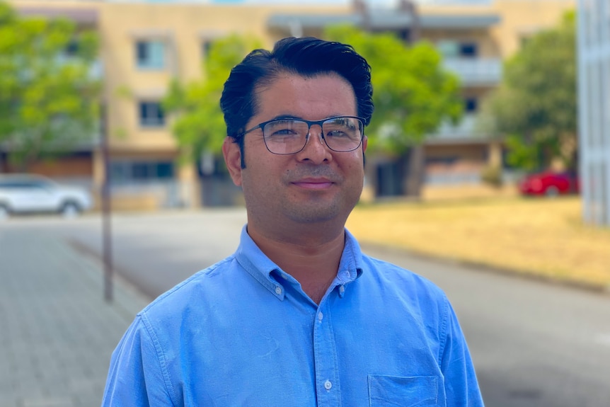 Assadullah Khurrami smiles while standing in front of a unit block wearing a bright blue shirt