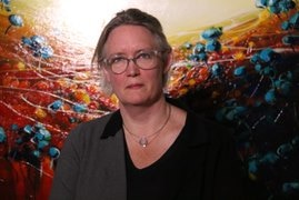 A woman wearing glasses stands in front of a painting of an object under a microscope.