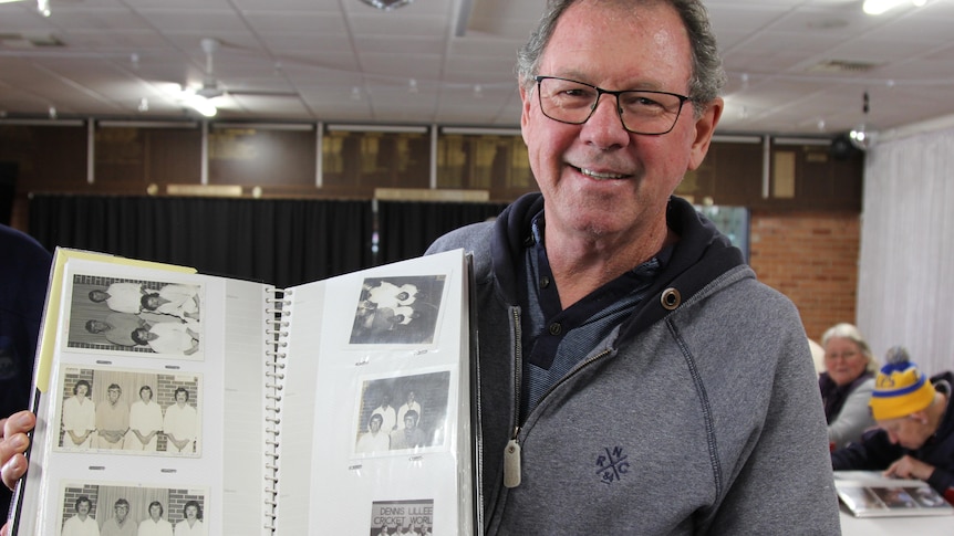 A man in a grey jumper holds a photo album with black and white photos of cricket players 