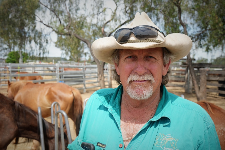 A man wearing an Akubra hat and a green shirt, standing outside with horses in the background.