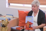A woman sits on a sofa in a school library and holds a book she wrote.