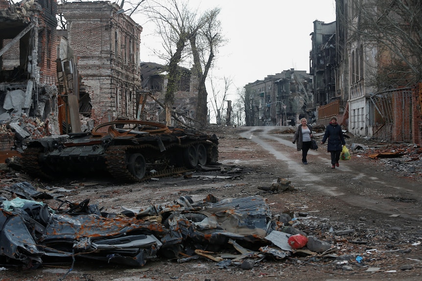 People walk past a destroyed tank and damaged buildings on a street littered with rubble and debris.