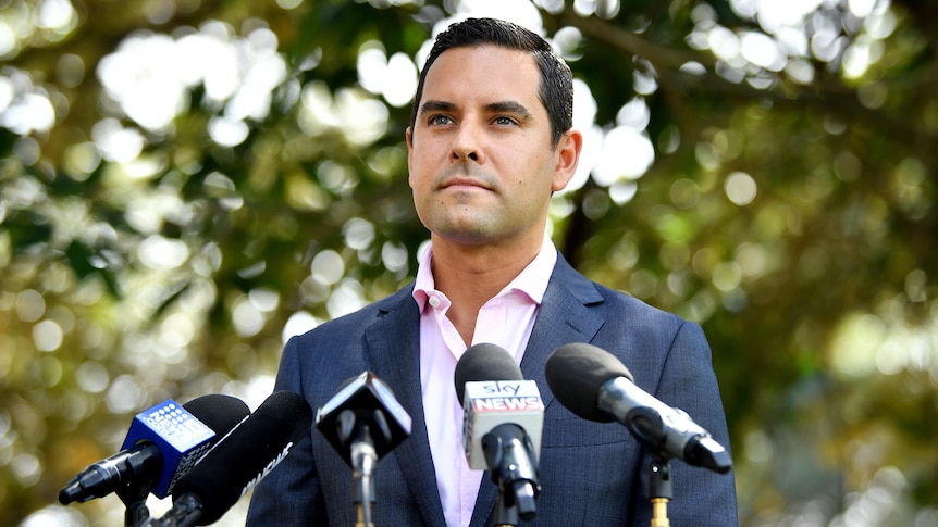 A young man standing outdoors behind microphones and talking to the media.