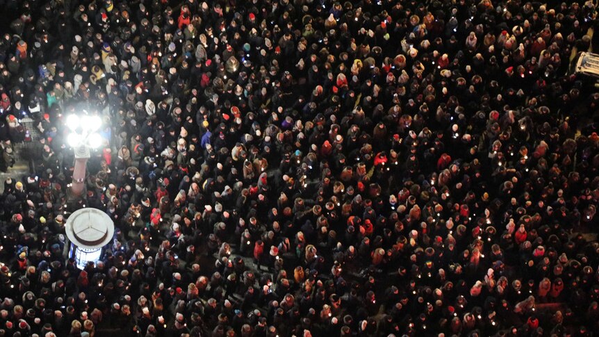 An aerial shot shows thousands of people gathered in the street at night. There are two lights poking out of the crowd.