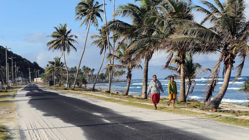 Two people walking along a sandy path beside palm trees that line a roadway