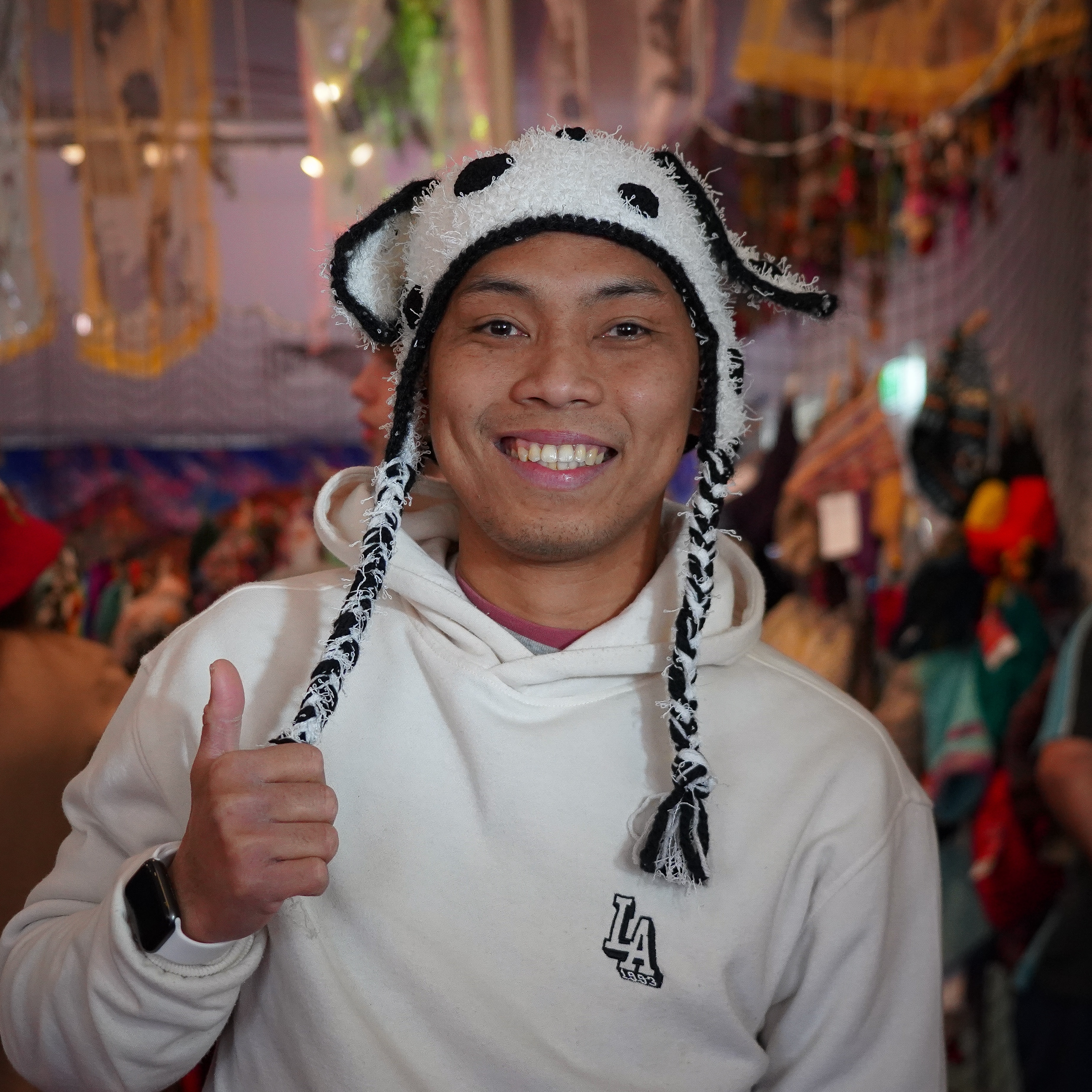 A man smiling at the camera with his thumbs up, he is wearing a knitted beanie that has black and white spots on it.