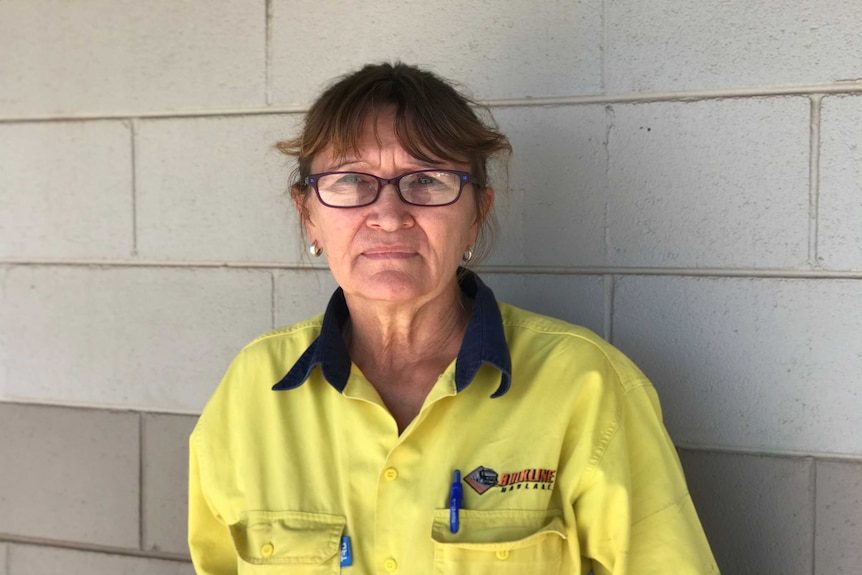 A lady stands against a white brick wall, wearing yellow hi-vis clothing and purple glasses