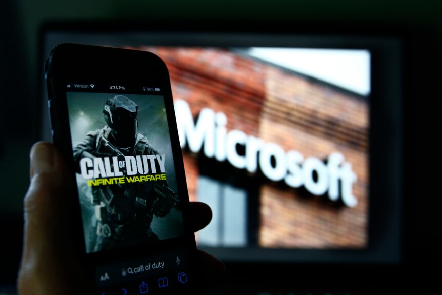 A person holds up a phone that shows a picture of Call of Duty in front of an image of a building with Microsoft signage. 