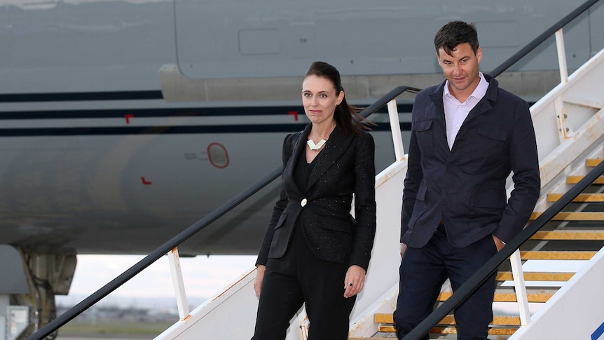 Jacinda Ardern exits an airplane down a set of stairs, followed by her partner Clarke Gayford. They are wearing dark suits.