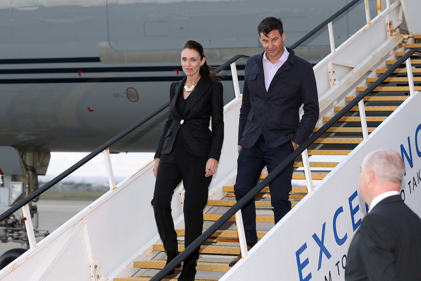 Jacinda Ardern exits an airplane down a set of stairs, followed by her partner Clarke Gayford. They are wearing dark suits.