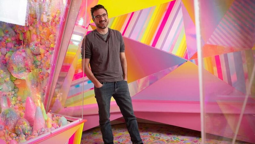 Man stands smiling with hands in jean pockets in front of a colour wall.