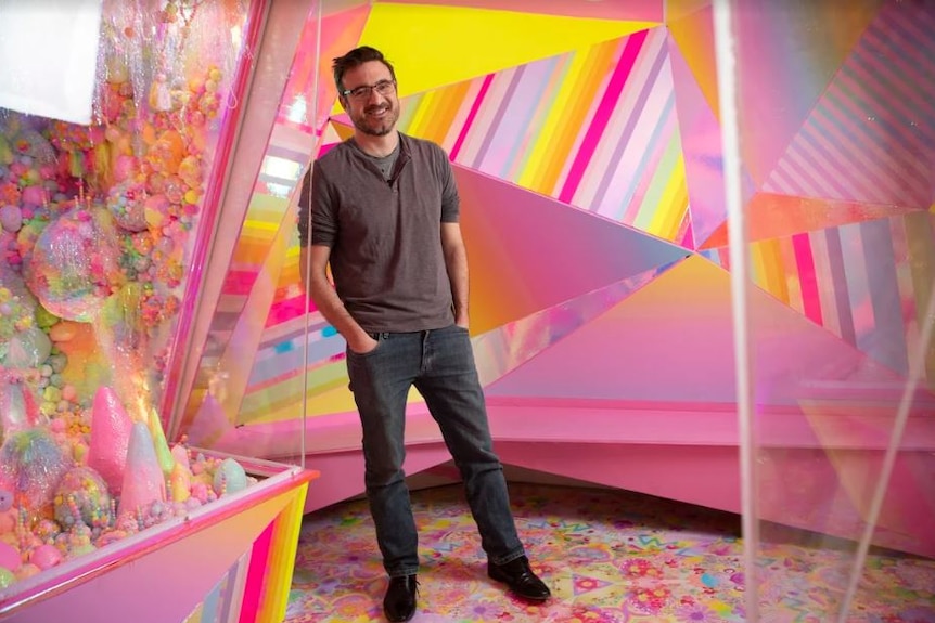 Man stands smiling with hands in jean pockets in front of a colour wall.
