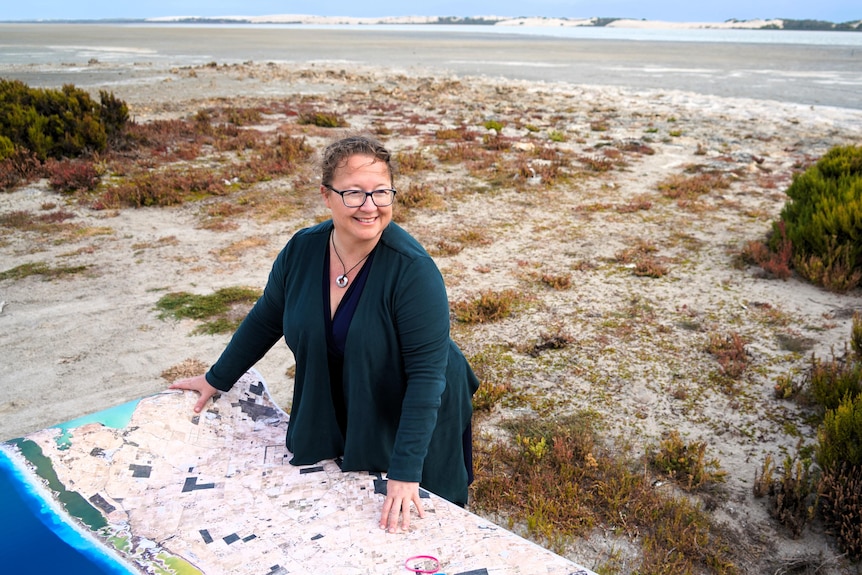 A woman stands with a map unrolled in front of her, a coastal lagoon with sand dunes behind her