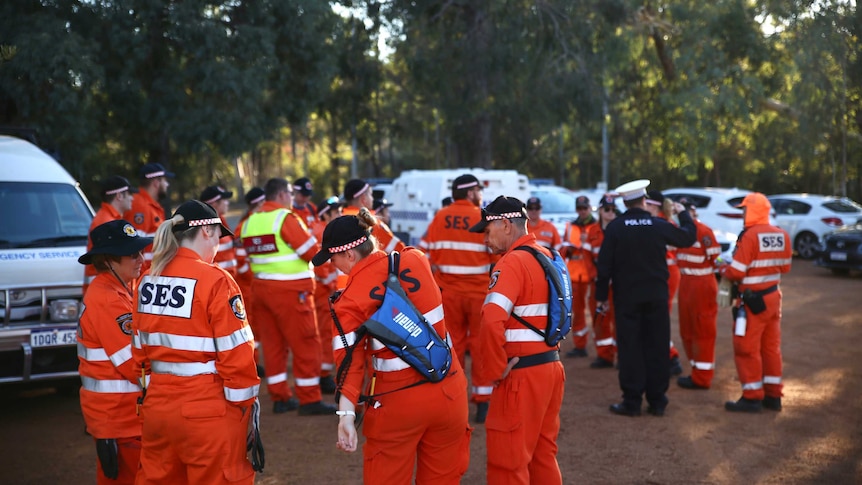A group of State Emergency Service volunteers stand wearing orange hi-vis clothing near cars and bushland.