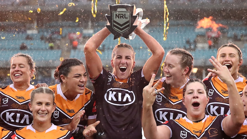 Ali Brigginshaw lifts the NRL trophy in the rain, with Brisbane Broncos teammates on either side of her.