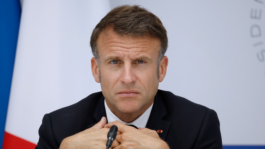 Emmanuel Macron sits with his hands crossed in a black suit looking stern