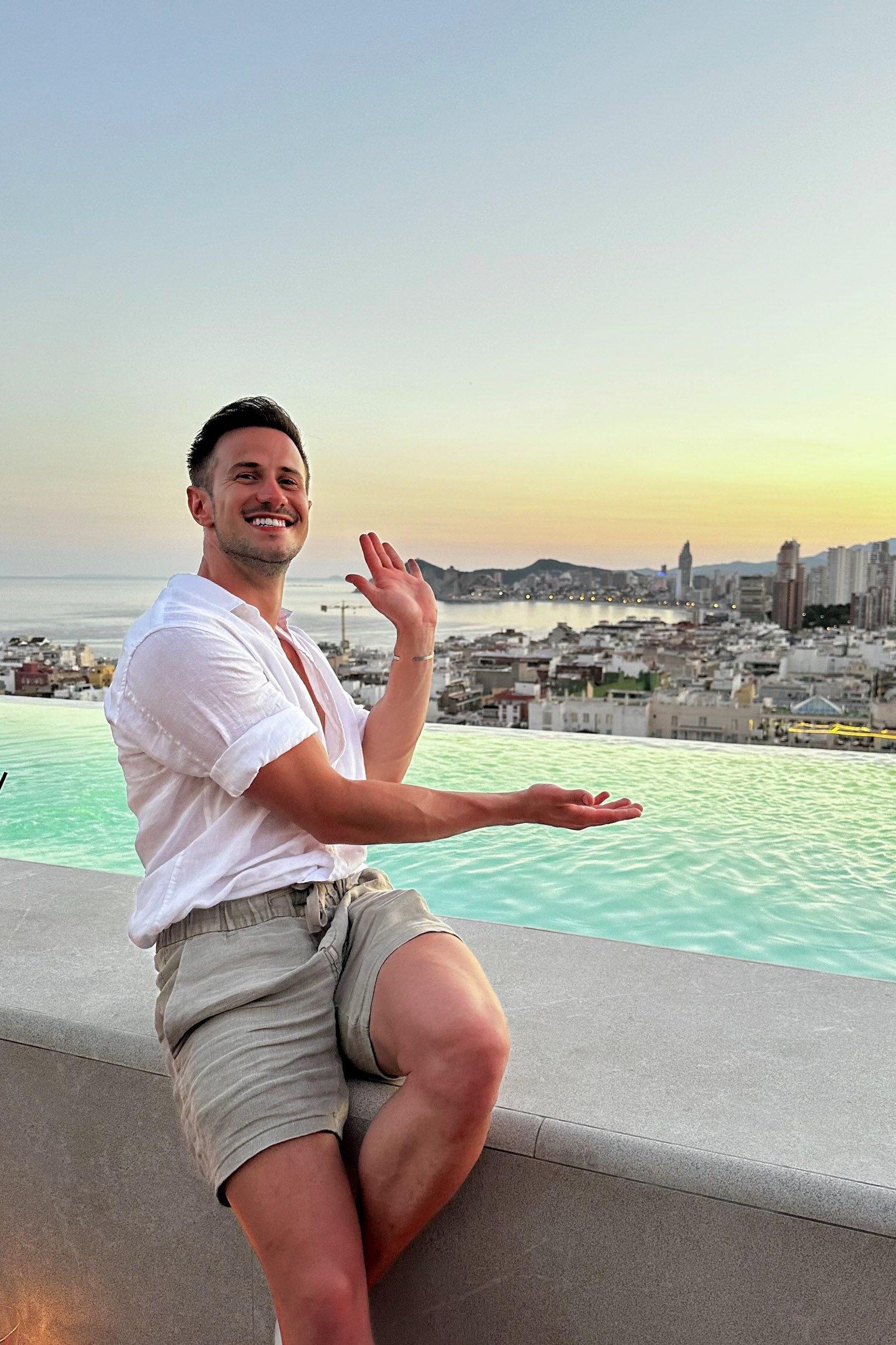 Scott, who has short brown hair, grins, sitting by the ocean during a stunning sunset.