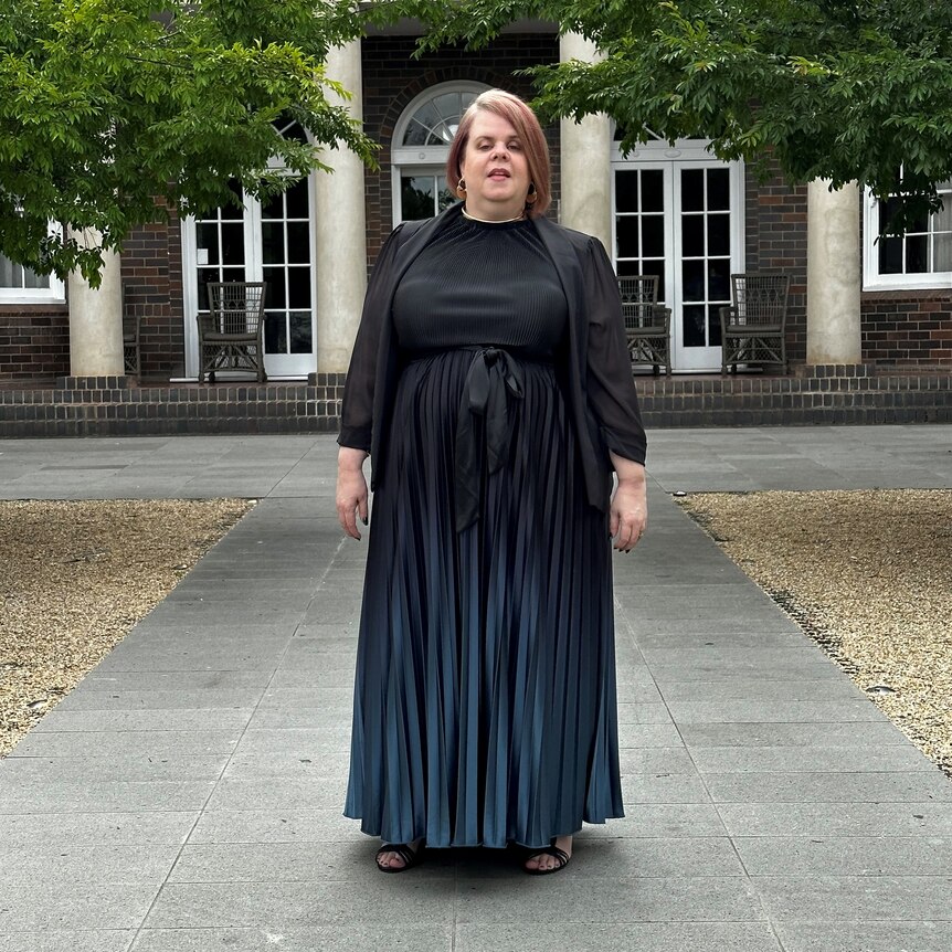Julee-ann Bell wears a black pleated dress and a jacket and stands in front of a columned building with tree leaves above her