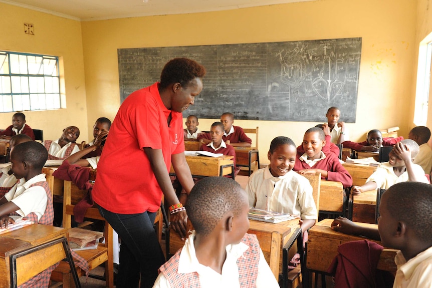 A teacher stands among young students sitting in a classroom.