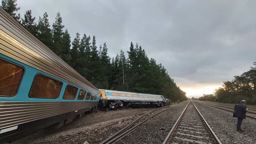 A train carriage lies on its side, a man stands nearby looking down the tracks.