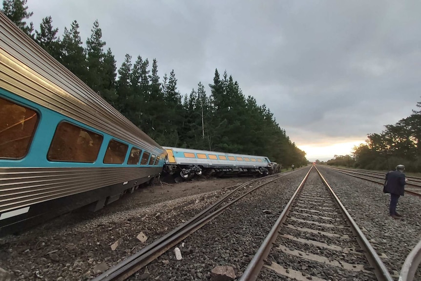 A train carriage lies on its side, a man stands nearby looking down the tracks.