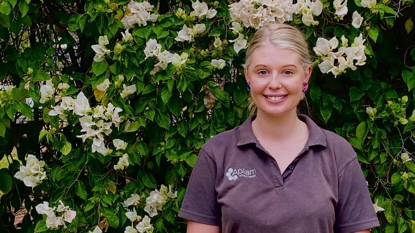 Monica, a young blonde woman and vet with outstanding HELP debt, stands smiling in front of a bush, wearing a polo shirt
