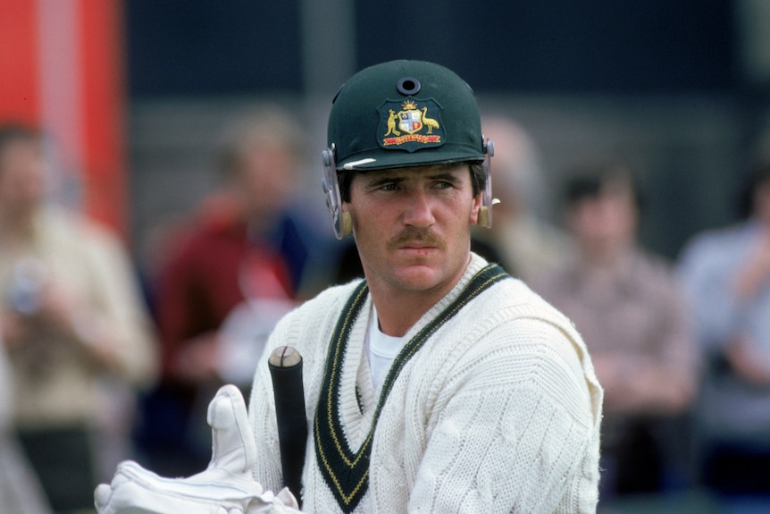 A mustached man with a green, grill-less helmet walks off the cricket field with his bat under his arm.