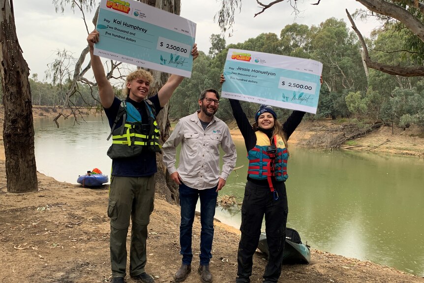 A tall teenage boy and young woman holding up large cheques, standing either side of a man wearing glasses, next to a river.