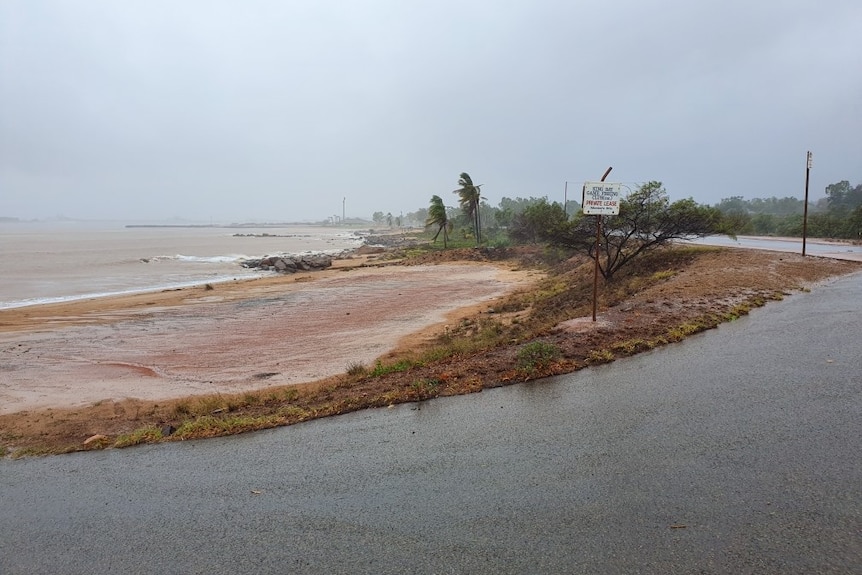 Palm trees blow in the wind and rain falls on the coast, with red dirt churned up.