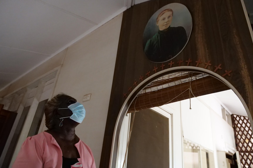 A woman gazes up at a painting of a woman in a green dress inside a dark, stained room
