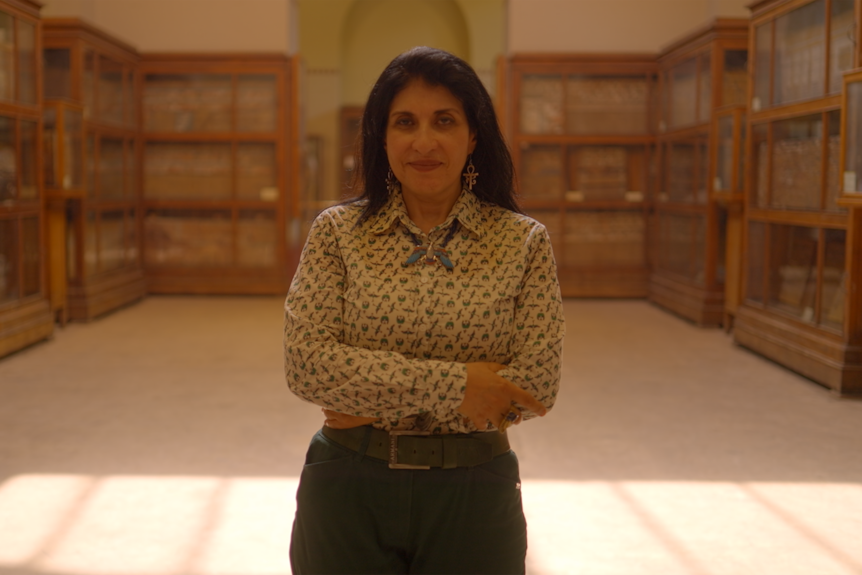 Egyptian woman with dark hair stands in a large museum room, her arms crossed. There are cupboards with glass doors behind her