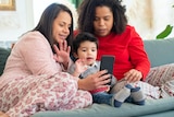 A family of two women and a child sit on the couch together in their pyjamas, looking at a phone.