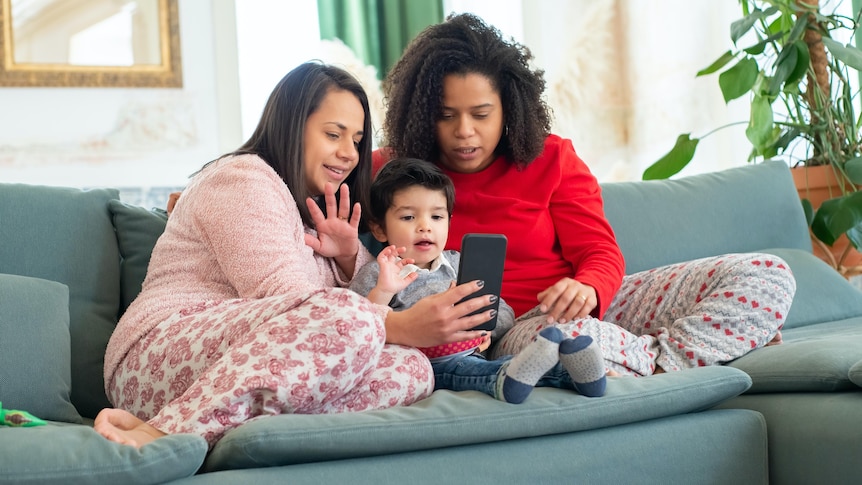 A family of two women and a child sit on the couch together in their pyjamas, looking at a phone.