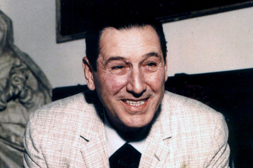 Man with dark hair smoking a cigarette and wearing a suit smiles at the camera.