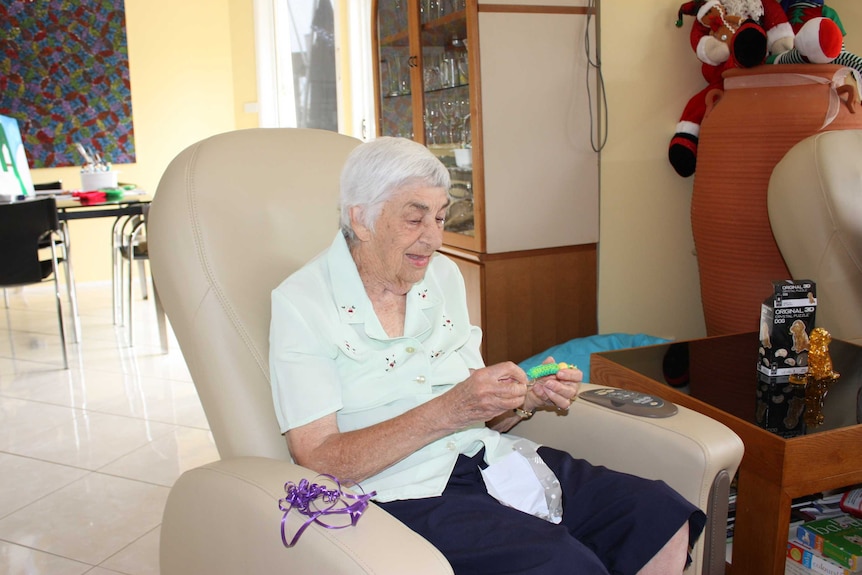 An elderly woman with white hair suits in a cream recliner in a lounge room, unwrapping a present.