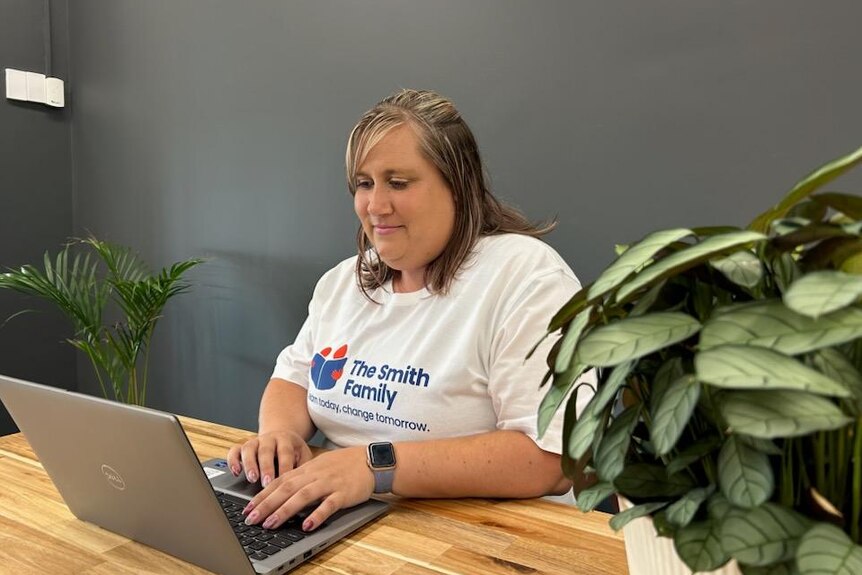 A woman in a shirt that reads 'Smith Familt' sits at a desk in front of her laptop.