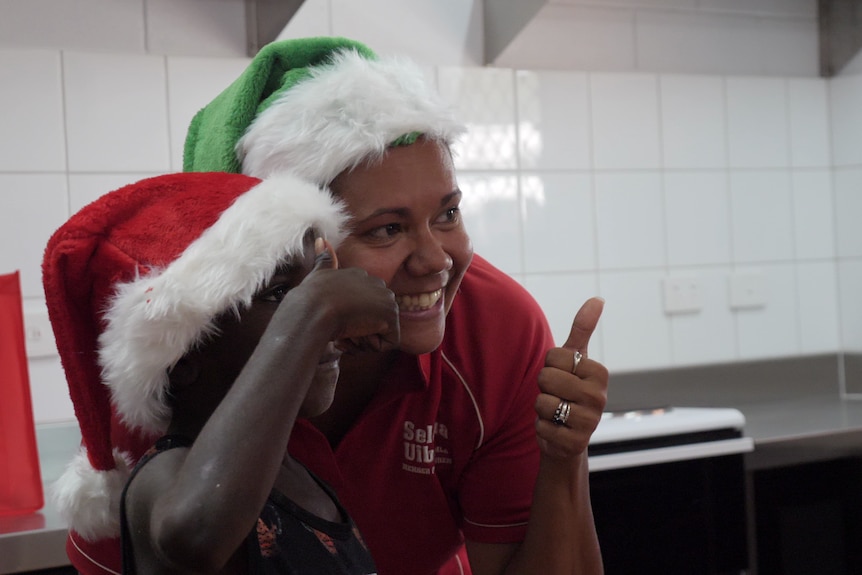 Selena Uibo smiles and gives the thumbs up while standing with a child also giving the thumbs up. Both are wearing santa hats.