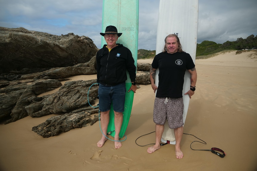 Two men in their 40s and 60s stand in front of their longboard on the beach, one aqua, one white.