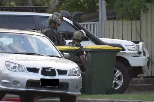 Two police officers talk to each other behind a car, outside a house where a siege is unfolding. 