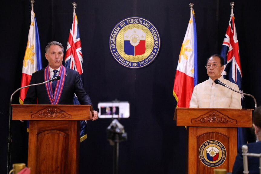Two men stand behind podiums at a press conference, flanked by flag poles and a country's national seal
