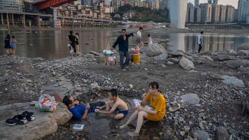 People sit in a shallow pool of water in a riverbed, with others swimming in the river in the background.
