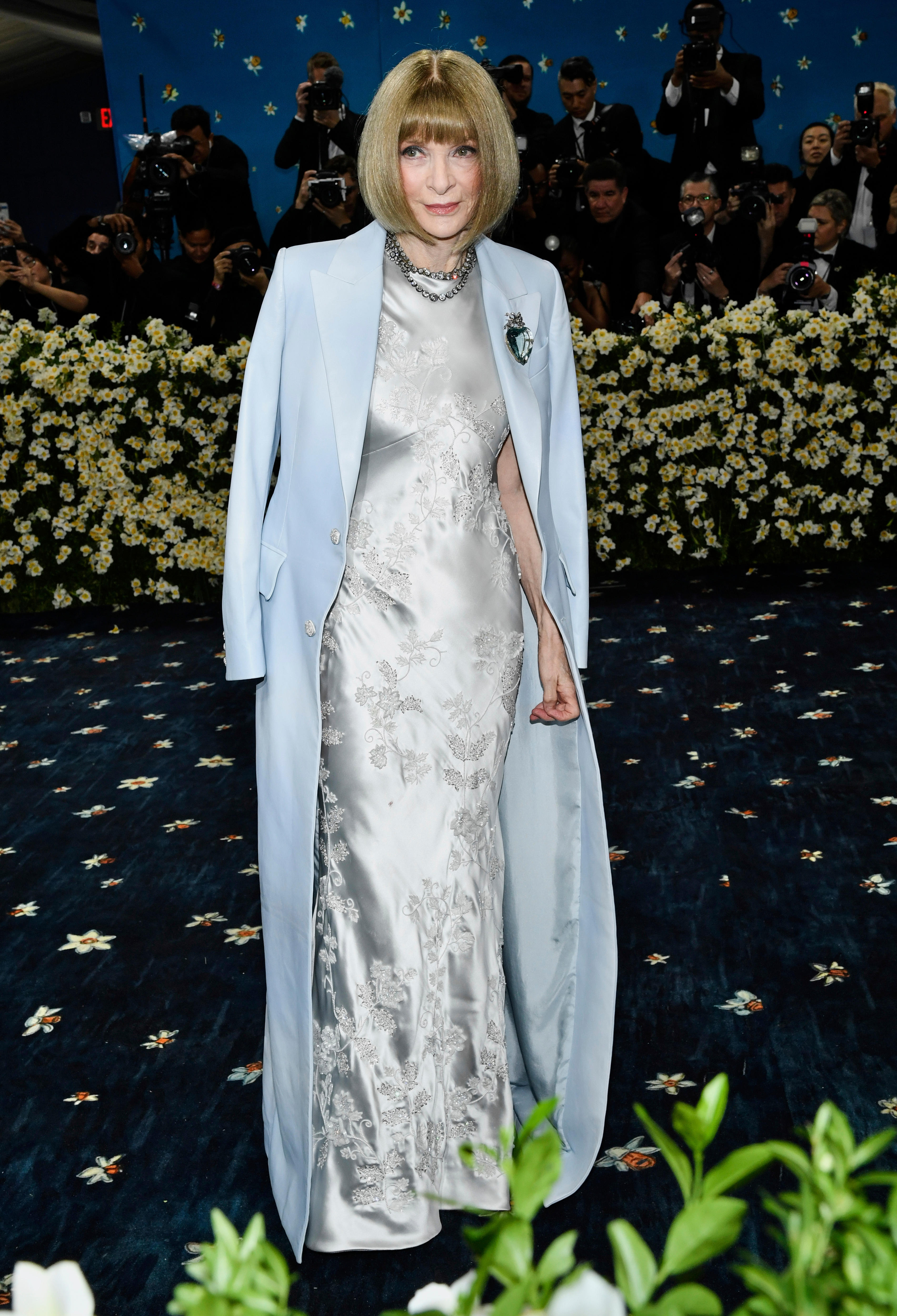 Anna Wintour wearing a long silvery dress with a floor-length baby blue suit jacked draped on her shoulders 