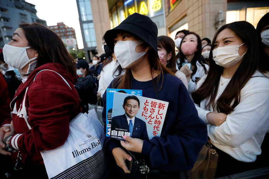 Three women wearing masks listen to political speeches.