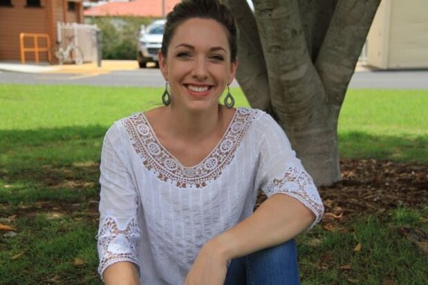 A young woman, wearing a white shirt and blue jeans, sits in a park and smiles.