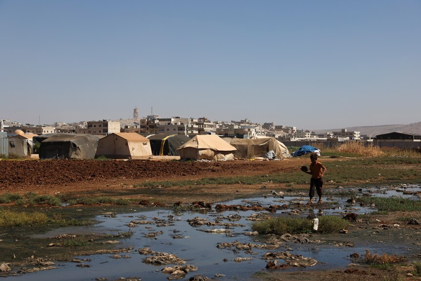 A child walks in a muddy stream next to a tent city. 